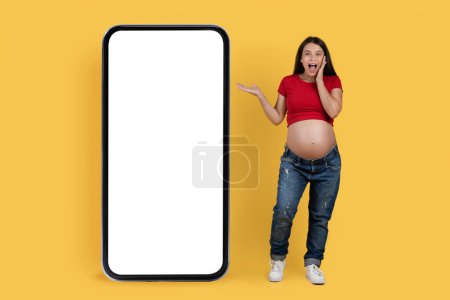 Foto de Increíble oferta. Mujer embarazada sorprendida señalando un gran teléfono inteligente en blanco con pantalla vacía blanca, sorprendida joven embarazada de pie aislada sobre fondo amarillo, collage, burla - Imagen libre de derechos