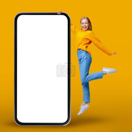Photo for Cheerful Young Woman Jumping Near Big Smartphone With Blank White Screen, Smiling Millennial Female Demonstrating Empty Mobile Phone While Having Fun Over Yellow Studio Background, Mockup - Royalty Free Image