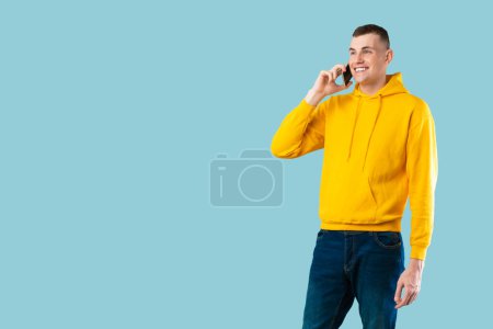 Foto de Siempre en contacto. Hombre caucásico casual hablando en el teléfono inteligente, mirando el espacio de copia, posando sobre fondo de estudio azul, bandera. Comunicación, concepto de llamada telefónica - Imagen libre de derechos