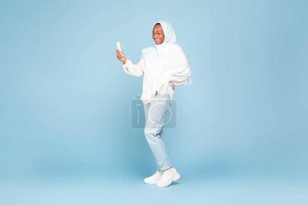 Photo for Happy african american woman in hijab using cellphone, standing on blue studio background, free space. Black lady chatting online, browsing social media, taking photo, full body length - Royalty Free Image