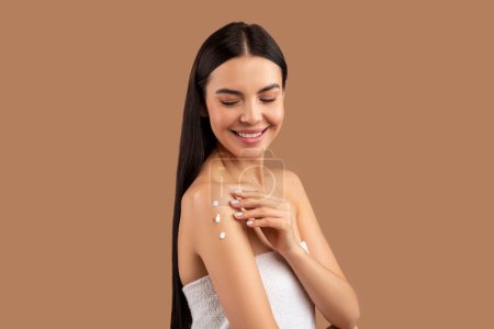 Photo for Smiling attractive shirtless long-haired brunette half-naked young woman applying body lotion or cream on shoulder, touching her smooth skin, isolated on beige studio background, copy space - Royalty Free Image