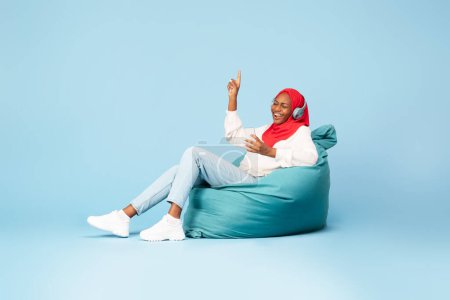 Foto de Mujer negra alegre en hijab con auriculares inalámbricos y teléfono inteligente escuchando música, disfrutando de la lista de reproducción, sentado en la silla de la bolsa de frijoles en el fondo del estudio azul, espacio para copiar - Imagen libre de derechos