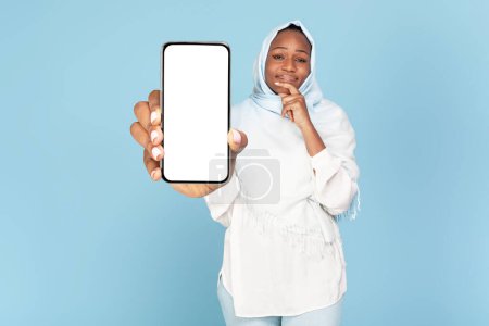 Téléchargez les photos : Femme musulmane noire réfléchie tenant un énorme smartphone avec écran vide à la main et en pensant, fond bleu, collage, maquette. Nouvelle application numérique, grande offre, annonce et vente - en image libre de droit