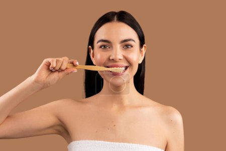 Téléchargez les photos : Portrait rapproché d'une jolie femme nue brune aux cheveux longs se brossant les dents avec une brosse à dents en bois biologique, nettoyant les dents isolées sur un fond de studio beige, concept de soins dentaires - en image libre de droit