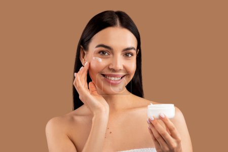 Photo for Beautiful naked brunette long-haired young woman applying facial cream on her skin and smiling, looking at camera and smiling, copy space, beige studio background, using eye face care cream - Royalty Free Image