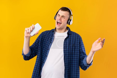 Photo for Joyful caucasian guy in wireless headphones singing song at smartphone as microphone, standing on blue background. Man enjoing favorite music - Royalty Free Image