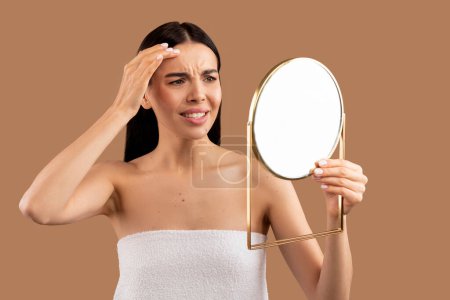 Foto de Mujer joven morena medio desnuda trastornada envuelta en toalla de baño mirando al espejo y tocando su cara sobre el fondo beige del estudio, sufriendo de problemas de piel. Acné, arrugas, piel opaca - Imagen libre de derechos