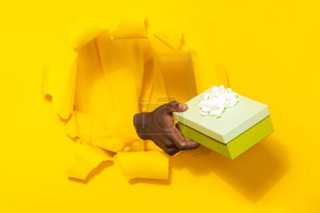 Téléchargez les photos : Homme afro-américain donnant boîte cadeau à travers fond de papier jaune déchiré. Main d'homme tenant présent dans l'emballage avec arc, gros plan - en image libre de droit
