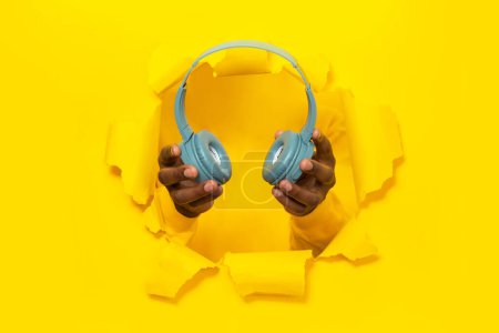 Foto de Hombre afroamericano sosteniendo auriculares inalámbricos modernos, rompiendo a través del agujero en el fondo de papel amarillo roto, primer plano. Tecnologías y nueva aplicación musical - Imagen libre de derechos