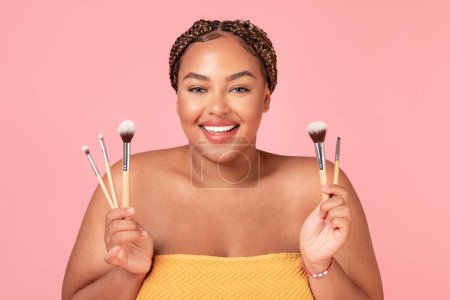 Photo for Make up concept. Portrait of beautiful black oversize lady holding set of makeup brushes, standing wrapped in towel over pink studio background, free space - Royalty Free Image