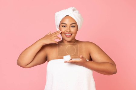 Téléchargez les photos : Concept de soins quotidiens. Portrait de femme afro-américaine en surpoids tenant un pot de crème et appliquant un hydratant sur la joue, posant à la caméra. Femme potelée noire portant une serviette blanche sur la tête - en image libre de droit