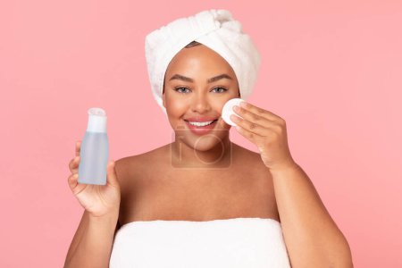 Téléchargez les photos : Portrait de belle femme noire obèse nettoyant sa peau avec du coton, tenant le flacon avec du toner et souriant à la caméra sur fond de studio rose, espace de copie - en image libre de droit