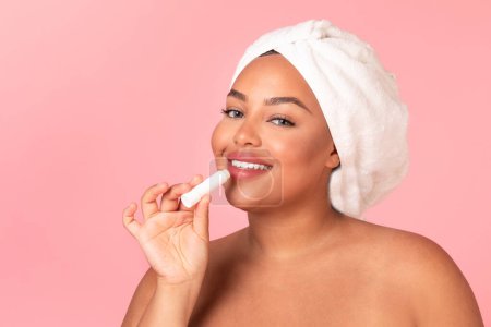 Photo for Chapped lips prevention. Beautiful black oversize woman using chapstick and smiling at camera, posing wrapped in towel and applying Llip balm over pink background, copy space - Royalty Free Image