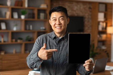 Téléchargez les photos : Souriant homme chinois d'âge moyen PDG montrer doigt à la tablette avec écran vide à l'intérieur du bureau à la maison. App et appareil pour les entreprises et le travail, recommandation et conseils nouveau site Web, covid-19 - en image libre de droit