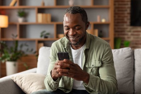 Foto de Nuevo dispositivo y aplicación. Retrato de hombre afroamericano de mediana edad sonriente lee el mensaje en el teléfono, mira el video, charlando en el interior de la sala de estar. Redes sociales, blog, búsqueda en internet en casa - Imagen libre de derechos