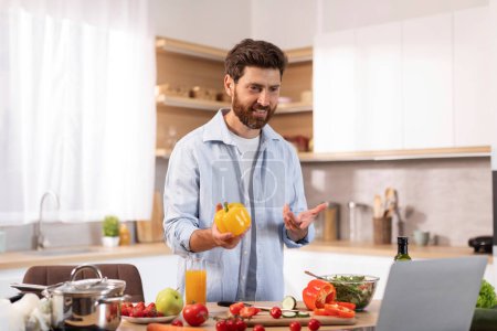 Photo for Glad mature caucasian guy with beard has video call, makes video at laptop, holds pepper in hands, prepares eat in modern kitchen interior. Homemade food, health care, meal blog with device at home - Royalty Free Image