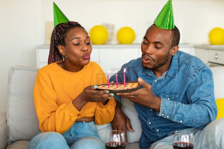 Foto de Emocionados cónyuges afroamericanos en sombreros festivos soplando velas encendidas en pastel de cumpleaños, celebrando b-day juntos, sentados en el sofá en casa y disfrutando de una pequeña fiesta - Imagen libre de derechos