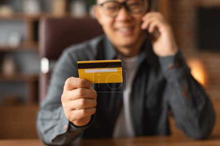 Photo for Happy adult asian male in glasses looks at credit card, sit at table, calls by phone in home office interior, cropped, blurred. Business remotely, investment and device, online shopping due covid-19 - Royalty Free Image