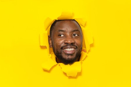 Photo for Positive african american man looking aside and smiling while tearing hole in yellow paper background, staring at copy space for your ad, panorama - Royalty Free Image