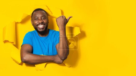 Téléchargez les photos : Homme afro-américain excité posant à travers un trou dans du papier déchiré, pointant le pouce vers l'espace libre, fond jaune, panorama. Publicité masculine grande offre et souriant à la caméra - en image libre de droit