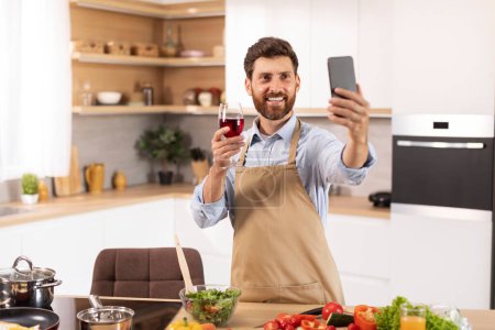 Photo for Smiling mature caucasian male with beard in apron hold glass of wine, looks at phone webcam, has video call, shooting video for blog in kitchen interior. Holiday celebration, congratulations at home - Royalty Free Image