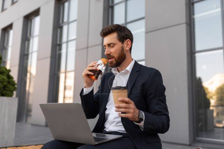 Photo for Serious young european man manager with beard in suit watch video on laptop with cup of coffee takeaway, eats croissant, sits on bench near building in city. Lunch outdoor, break from work, business - Royalty Free Image