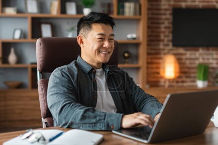 Cheerful attractive mature asian businessman look at computer at table, has video call at home office interior. Work, business remotely and freelance, meeting with technology during covid-19 pandemic