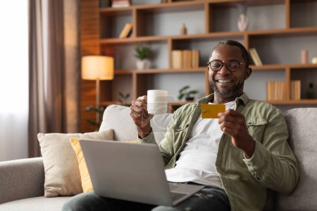 Téléchargez les photos : Joyeux homme afro-américain d'âge moyen dans des lunettes avec ordinateur portable bénéficie d'une tasse de café et montre carte de crédit, utiliser la finance bancaire dans le salon intérieur. Achats en ligne, vente, nouvelle normale et gadget - en image libre de droit