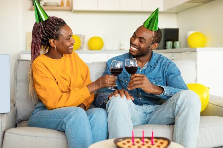 Photo for Loving black spouses clinking glasses of wine, celebrating birthday together at home, sitting on sofa and smiling at each other. Family enjoying b-day party - Royalty Free Image
