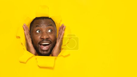 Foto de Wow ofrecen concepto de publicidad. Hombre negro emocional mirando el espacio libre y tocando las mejillas, posando en un fondo de papel amarillo roto, panorama - Imagen libre de derechos