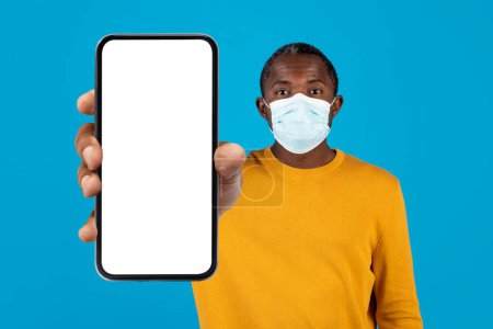 Foto de Hombre negro de mediana edad en máscara médica protectora en la cara que muestra el teléfono celular con pantalla blanca en blanco, que muestra la aplicación con el mapa de seguimiento de brotes de coronavirus, fondo azul del estudio, maqueta - Imagen libre de derechos