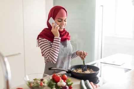 Photo for Cheerful young muslim woman in hijab housewife cooking tasty dinner at kitchen, mixing food in frying pot and talking on phone, having chat with her friend while preparing meal, copy space - Royalty Free Image