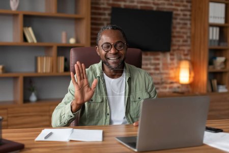 Téléchargez les photos : Souriant homme d'affaires afro-américain d'âge moyen dans des lunettes agitant la main à l'ordinateur portable webcam, a réunion à l'intérieur du bureau à la maison. Salut, salut et bonjour, appel vidéo, travail d'étude et affaires à distance - en image libre de droit