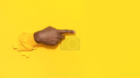 Foto de Mano masculina afroamericana apuntando a un lado en el texto o su diseño con el dedo índice, haciendo un gesto a través del fondo de papel amarillo rasgado, primer plano, espacio libre - Imagen libre de derechos