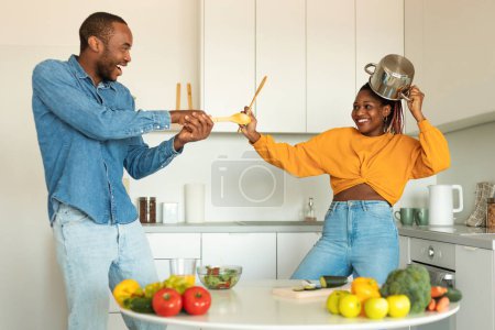 Téléchargez les photos : Amusement domestique. Joyeux couple noir ludique se battant dans la cuisine, en utilisant des spatules comme armes, conjoints heureux tromper ensemble à la maison, profiter du temps avec l'autre - en image libre de droit