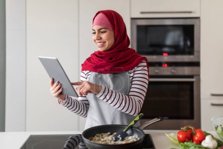 Foto de Alegre mujer joven de Oriente Medio en hijab comprobar la receta en Internet mientras prepara comida saludable para su familia en casa, la celebración de la tableta digital, la lectura de blog culinario, interior de la cocina moderna - Imagen libre de derechos