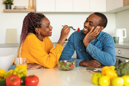 Photo for Loving black woman feeding her boyfriend with fresh salad, eating in cozy kitchen together, having lunch at home and smiling to each other, free space - Royalty Free Image