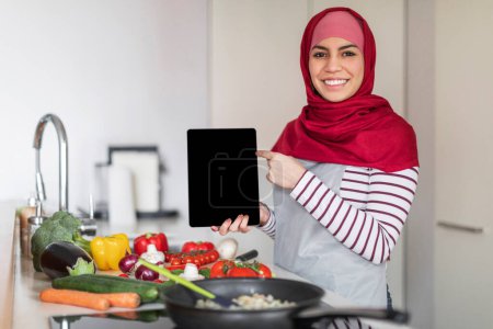 Téléchargez les photos : Jolie jeune femme musulmane du Moyen-Orient portant hijab cuisiner de délicieux aliments sains à la maison, montrant tablette numérique avec écran noir vierge, recommandant belle application de cuisine, espace de copie, maquette - en image libre de droit