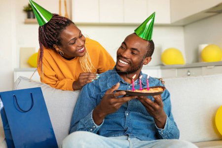 Foto de Amante esposa negra haciendo sorpresa para su marido, hombre feliz sosteniendo pastel de cumpleaños mientras está sentado en el sofá, espacio libre. Familia en sombreros de fiesta celebrando cumpleaños en casa - Imagen libre de derechos