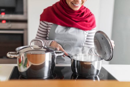 Photo for Unrecognizable middle eastern muslim housewife woman wearing hijab cooking meal, checking pot on electric stove, enjoying cooking at home, kitchen interior, cropped, copy space - Royalty Free Image