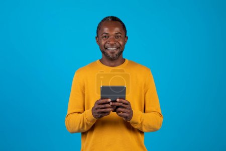 Photo for Positive attractive grey-haired middle aged black man in yellow using brand new digital pad and smiling at camera on blue studio background, surfing on Internet, using mobile app, copy space - Royalty Free Image