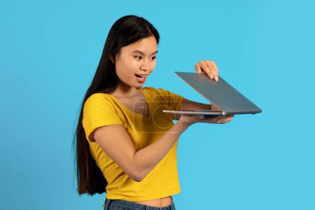 Téléchargez les photos : Joyeux curieux jolie millénaire étudiante chinoise en t-shirt jaune ouvre ordinateur portable, isolé sur fond bleu, studio. Surprise, technologie pour le travail et l'étude, grande annonce et offre, émotions - en image libre de droit