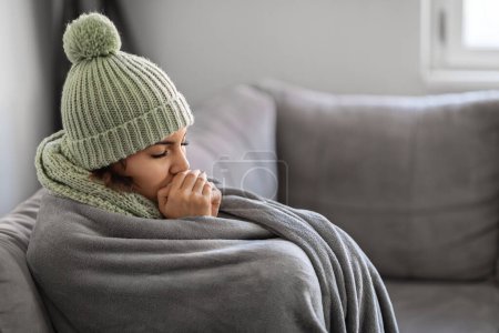 Foto de La mujer que se congela que calienta las manos con la respiración mientras que se sienta en el sofá en el país, mujer joven que usa el sombrero y la bufanda tejidos que se sienten fríos en el interior, señora cubierta en manta que sufre problemas de calefacción en plano - Imagen libre de derechos