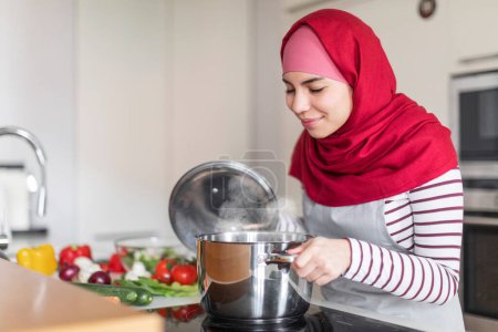 Foto de Alegre mujer musulmana de Oriente Medio ama de casa que usa comida de cocina hijab, comprobando la olla en la estufa eléctrica, oliendo con los ojos cerrados, disfrutando de cocinar en casa, interior de la cocina, recortado, espacio de copia - Imagen libre de derechos