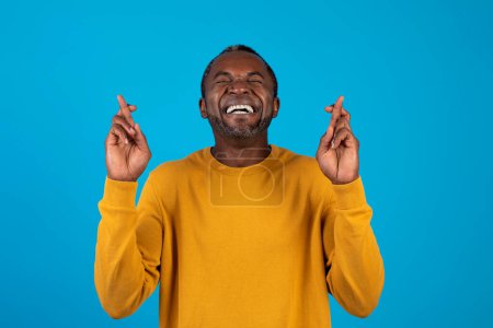 Photo for Making Wish. Superstitious emotional middle aged black man in yellow sweater with closed eyes keeping his fingers crossed, praying for luck, isolated on blue studio background, copy space - Royalty Free Image
