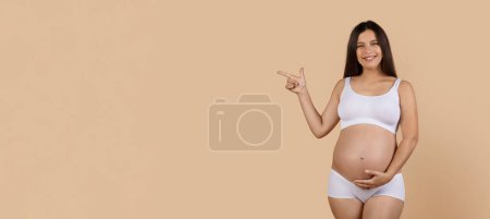 Téléchargez les photos : Regarde là. Femme enceinte souriante en sous-vêtements pointant du doigt à l'espace de copie, jeune femme enceinte heureuse indiquant un endroit gratuit pour la publicité tout en se tenant debout sur fond de studio beige, Panorama - en image libre de droit