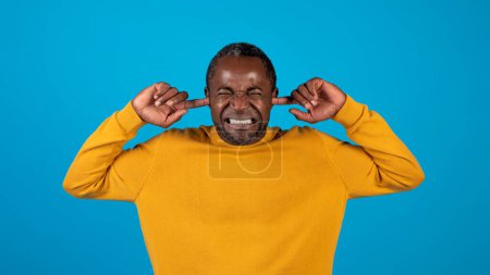 Téléchargez les photos : Homme mûr afro-américain bouchant les oreilles avec les doigts. Homme en jaune couvrant les oreilles avec un visage en colère grave, fronçant les sourcils, criant, grincement de dents. Portrait, plan studio, concept de colère, fond bleu - en image libre de droit