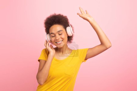 Photo for Satisfied relaxed young african american curly woman in casual, wireless headphones, dancing, enjoy music, isolated on pink background, studio. Fun alone at spare time, audio app and gadget for rest - Royalty Free Image