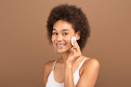 Photo for Cheerful millennial african american curly lady in white top removes skin with cotton pad isolated on brown background, studio. Cosmetics, moistening, beauty care at home, daily treatments and spa day - Royalty Free Image