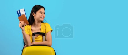 Foto de Feliz millennial mujer coreana turista en camiseta amarilla con maleta mostrar pasaporte, entradas, mira el espacio vacío, aislado sobre fondo azul, estudio, panorama. Viajes, vacaciones, viajes, anuncios y ofertas - Imagen libre de derechos